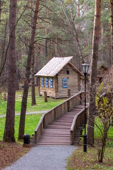 Wooden house with a manor house in the forest. The house has painted Windows. A wooden bridge leads to the house across the river. 