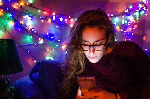 Night photography of lifestyle interior in the bedroom with a Caucasian and blonde girl looking at her mobile and with colored led lights on the head of the bed