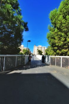 Elche, Alicante, Spain- October 7, 2020: People crossing a pedestrian bridge in the morning in Elche. Beautiful pine trees in both sides.