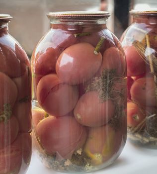 Home preservation: large glass jars with red ripe pickled tomatoes, closed with metal lids, stand on the windowsill. Front view, close-up.