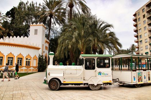 Elche, Alicante, Spain- February 19, 2019: Guided tour on Tourist train through the streets and palm groves in winter under blue sky. Tourist Office in the background.