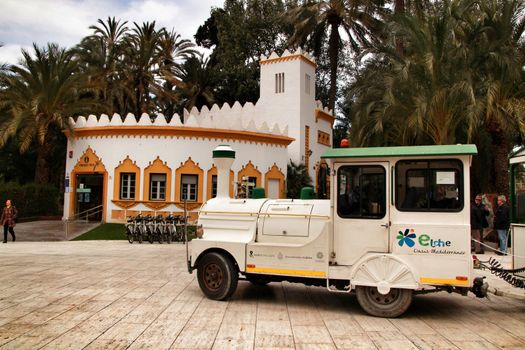 Elche, Alicante, Spain- February 19, 2019: Guided tour on Tourist train through the streets and palm groves in winter under blue sky. Tourist Office in the background.