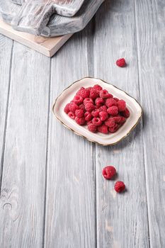 Raspberry fruits in plate near to old cutting boards, healthy pile of summer berries on grey wooden background, angle view