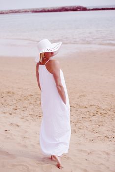 Woman walking on beach in long white dress and hat