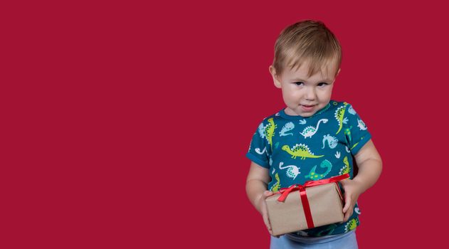 A little boy tries to unpack a New Year's gift with enthusiasm and excitement looking at the camera smiling on a red background