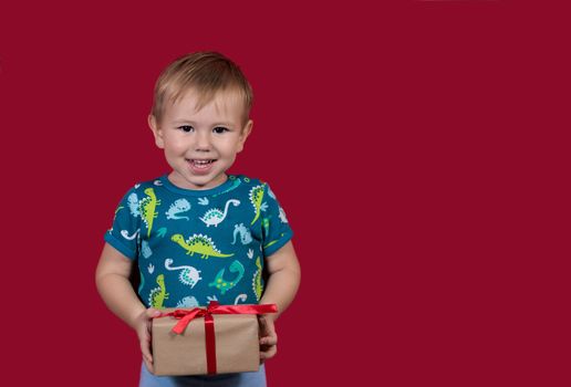 A little boy tries to unpack a New Year's gift with enthusiasm and excitement looking at the camera smiling on a red background