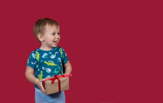 A little boy tries to unpack a New Year's gift with enthusiasm and excitement looking aside smiling on a red background