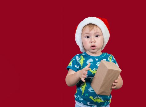 A little boy wearing a Santa hat tries to unpack a New Year's gift with enthusiasm and excitement on a red background