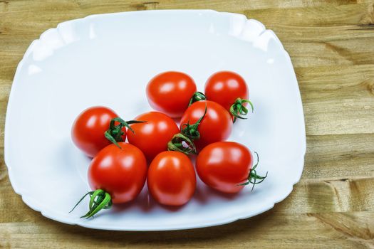 On a wooden table is a white plate with red small tomatoes