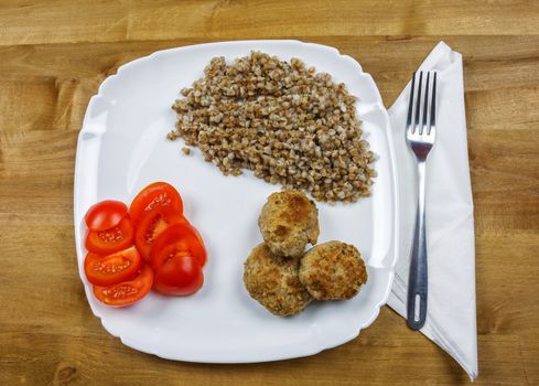 On a square plate is buckwheat porridge with meat cutlets and chopped tomato