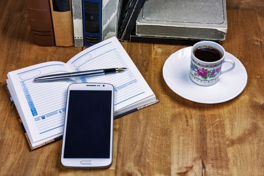 On the wooden surface are books, a cup of coffee, a diary with a pen and a smartphone