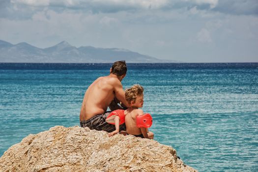 A man and a little boy are sitting on the stone near the sea