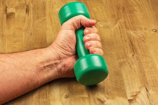 A man's hand holds a green dumbbell against the background of a wooden surface