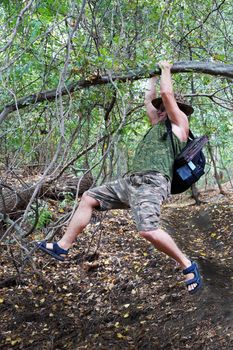 a man with a backpack climbs over a ravine hanging on his hands on a tree.