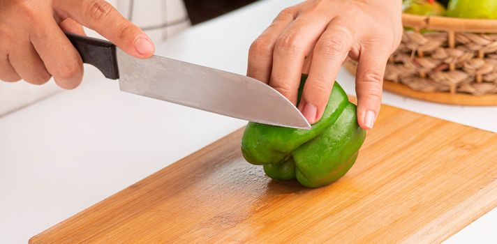Woman cutting Fresh Green Bell Pepper into pieces with knife on wooden chopping board.