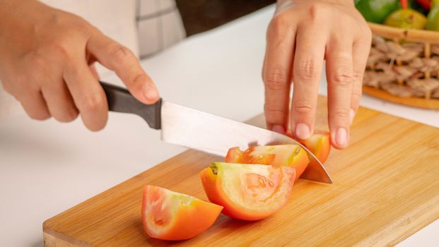 Woman cutting Fresh Tomato into pieces with knife on wooden chopping board.