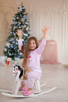 A beautiful little girl in pink pajamas rejoices in a wooden rocking horse, a gift from Santa for Christmas.