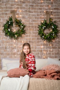 Little girl in red pajamas on the bed in a cozy Christmas interior.