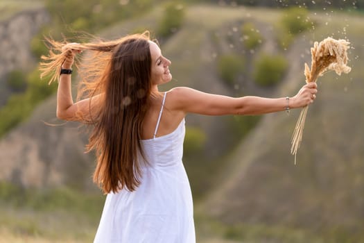 Beautiful tender girl in a white sundress walks at sunset in a field with a spikelet bouquet