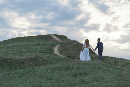 A couple in love, a pregnant wife in a white outfit are walking in a hilly field. Overcast weather,