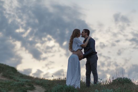 A couple in love, a pregnant wife in a white outfit are walking in a hilly field. Overcast weather,