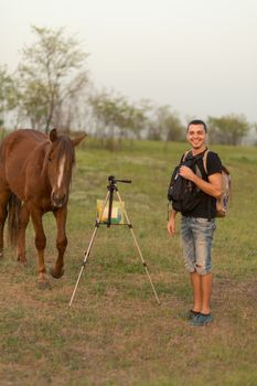 A guy with a tripod for a photo video shooting in a meadow with a horse. The horse is interested.