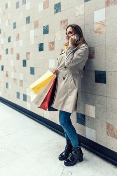 Adult woman wearing a beige raincoat and yellow scarf talking on her mobile phone while holding shopping colorful bags.