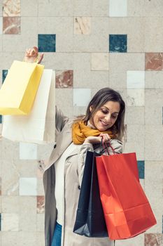 Adult funny woman wearing a beige raincoat and yellow scarf holding shopping colorful bags