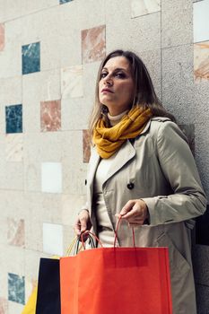 Adult woman wearing a beige raincoat and yellow scarf holding shopping colorful bags.
