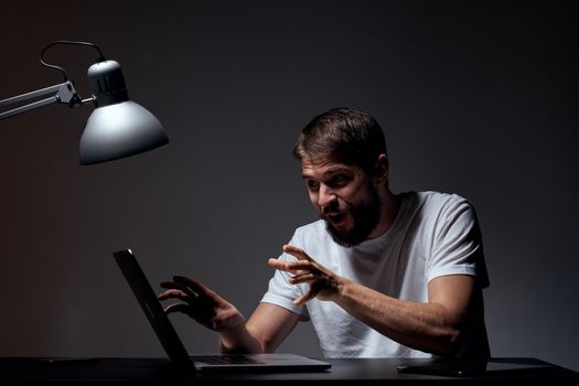 man with laptop sitting at table dark background workplace office emotions lamp gesturing with hands model cropped view. High quality photo