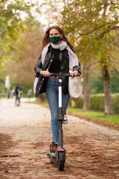 Casual caucasian teenager wearing protective face mask riding urban electric scooter in city park during covid pandemic. Urban mobility concept.