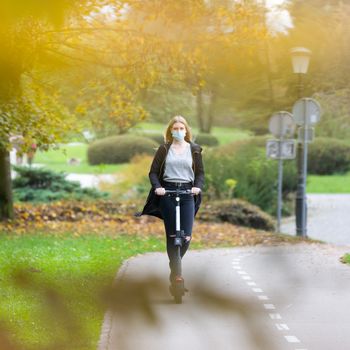 Casual caucasian teenager wearing protective face mask riding urban electric scooter in city park during covid pandemic. Urban mobility concept.