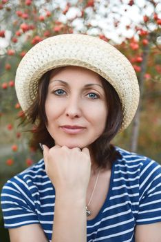 Image of beautiful middle-aged woman wearing straw hat laughing and looking at camera while walking in summer park