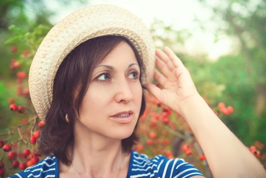 Image of beautiful middle-aged woman wearing straw hat laughing and looking at camera while walking in summer park