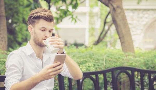 Business young handsome man drink cup of coffee using mobile phone sitting in the park, communication concept.