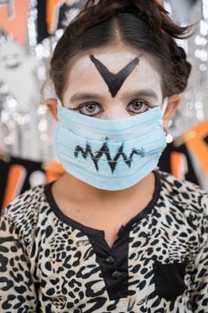 Kid in Halloween witch dress and scary face with medical mask looking into camera - halloween celebration during pandemic