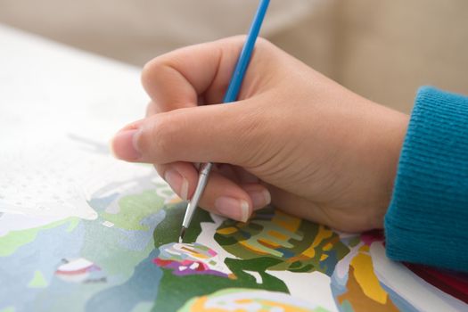 4 - Young and youthful female hand with natural nails is shown in great detail holding a blue paint brush and painting by numbers. A popular hobby.