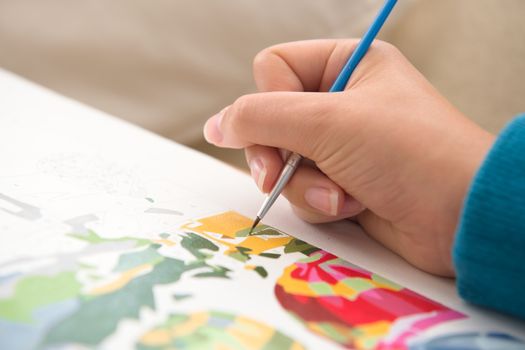 8 - Detailed close up of thin paint brush being used by a young female to paint by numbers. Multiple colors and vivid blues contrast the warm skin.