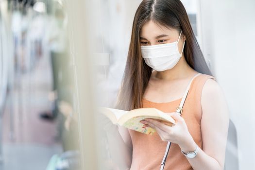 Young Asian woman passenger wearing surgical mask and reading the book in subway train when traveling in big city at Covid19 outbreak, Infection and Pandemic concept