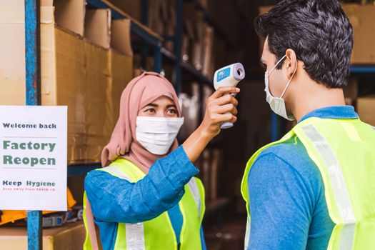 Muslim woman worker uses Medical Digital Infrared Thermometer measure temperature to Indian man worker with safety clothes before start to work after warehouse reopen again, new normal concept