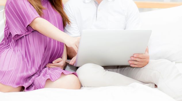 Beautiful pregnant family with laptop computer on bedroom, husband and wife notebook relax together, couple and family concept.