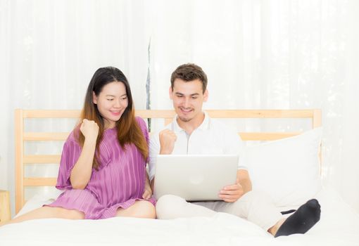 Beautiful pregnant family with laptop computer on bedroom, husband and wife notebook relax together, couple and family and success concept.