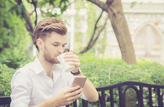 Business young handsome man drink cup of coffee using mobile phone sitting in the park, communication concept.