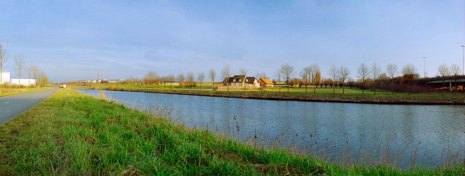 Panoramic view of a river with trees in warm winter light before sunset with a cool blue couldless sky with a lot of green grass and factorys in the background