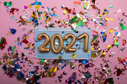 Gold numbers 2021 on face mask, pink background and confetti. Happy new pandemic year