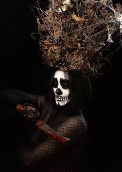 woman in black clothes and skull makeup, a crown of dry branches and flowers on her head, black background, Halloween carnival costume, holds a knife with streaks of blood in his hands