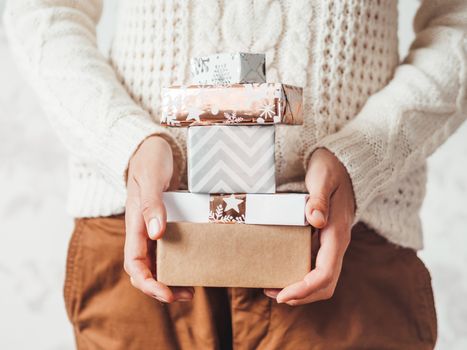 Woman in cable-knit white sweater with Scandinavian pattern and brown chinos trousers with leather belt. Stack of Christmas presents in hands. Casual outfit for New Year celebration.