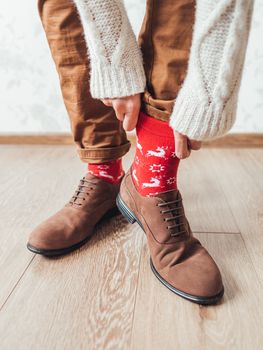 Young man pulls up leg of his chinos trousers to show bright red socks with reindeers on them. Scandinavian pattern. Winter holiday spirit. Casual outfit for New Year and Christmas celebration.