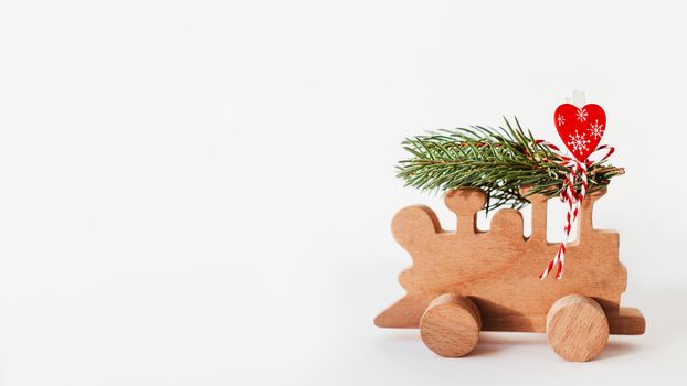 Wooden train with tied fir tree branches and red heart decoration. Cute symbol of Christmas tree brought home for New Year celebration. Winter holiday spirit. Copy space on white background.