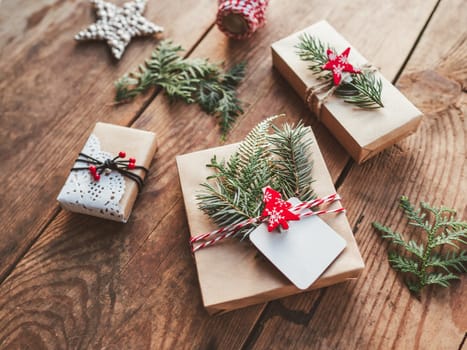 Christmas DIY presents wrapped in craft paper with fir tree twigs. Red decorations in shape of Christmas tree, stars. New Year gifts on wooden background. Winter holiday spirit.